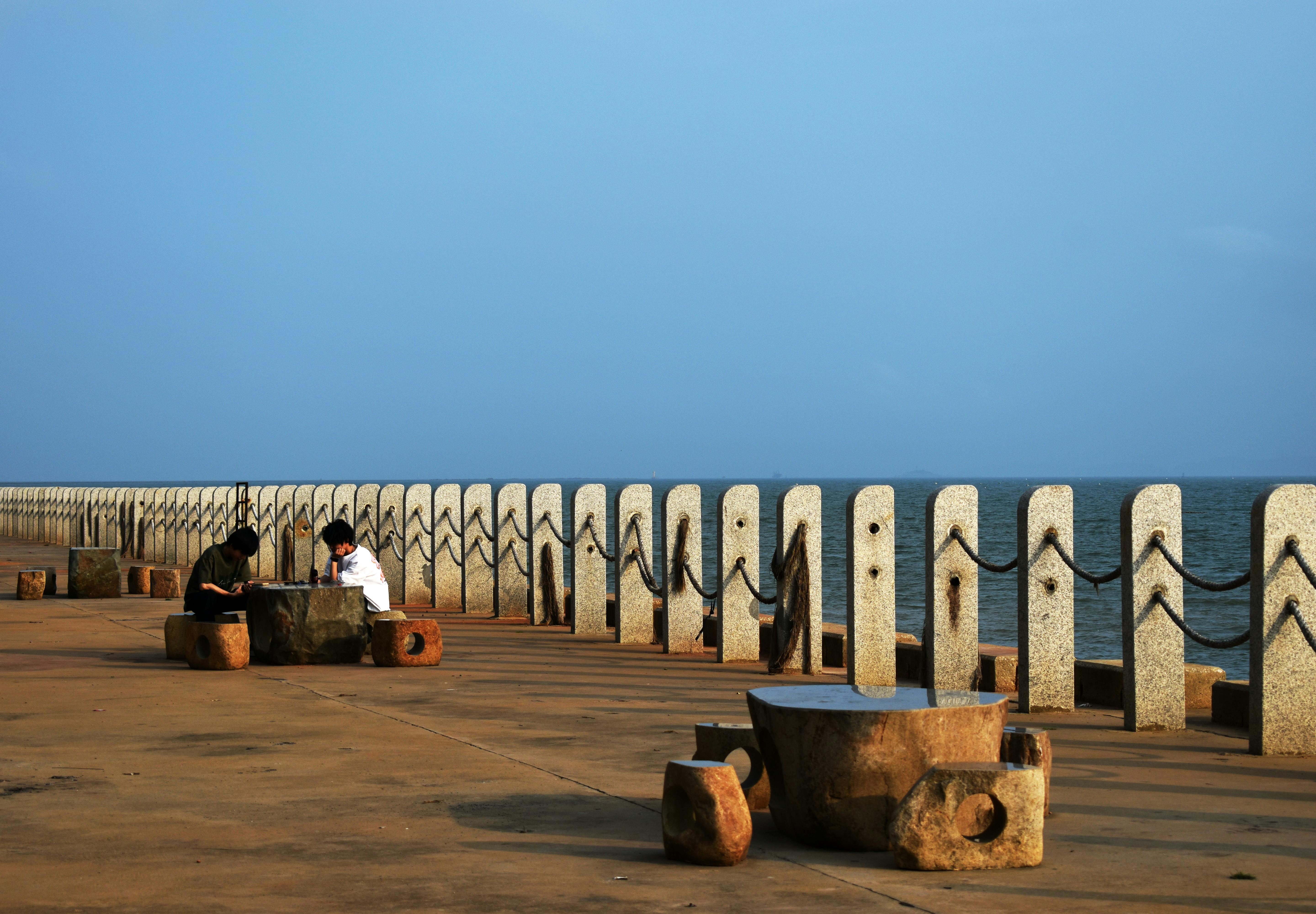 Two people enjoy a peaceful moment by the seaside on a sunny day, sitting by a concrete barrier.
