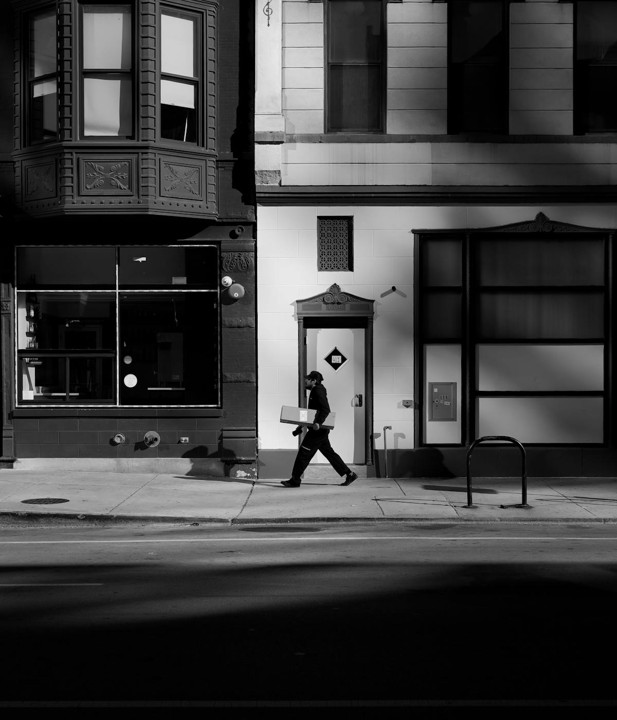 Black and white photograph capturing a person in a cap and dark clothing walking on a sidewalk while carrying a box.