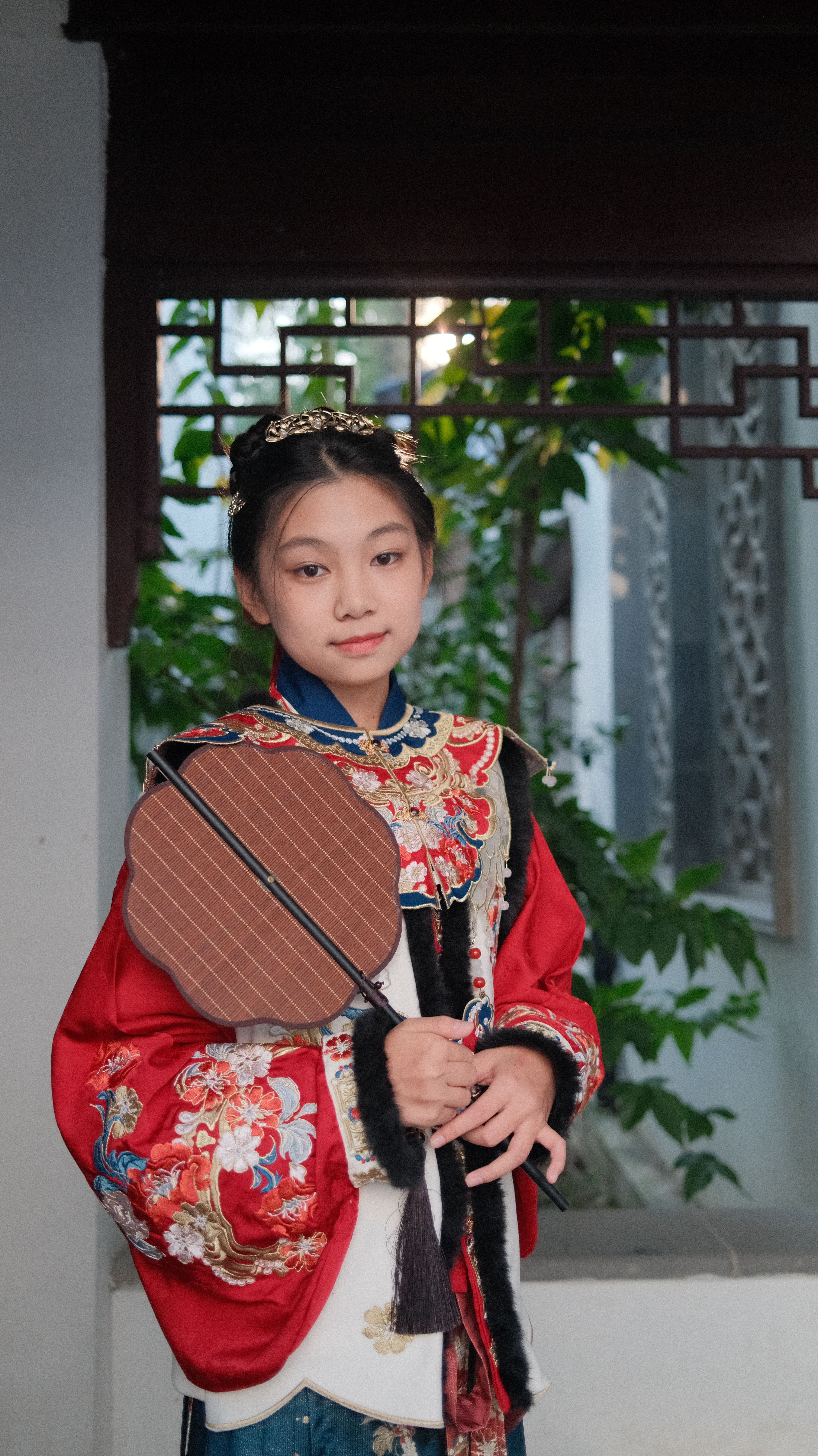 A young girl wearing traditional Chinese clothing holds a wooden fan indoors.