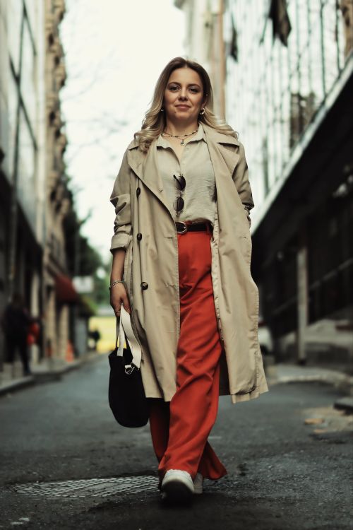 Free Stylish woman in trench coat and red pants walking confidently down a city street. Stock Photo