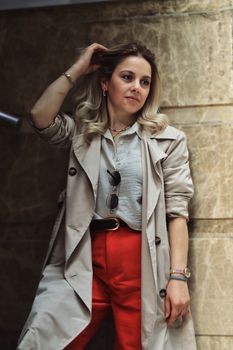 Fashionable woman posing indoors wearing a trench coat and red pants.