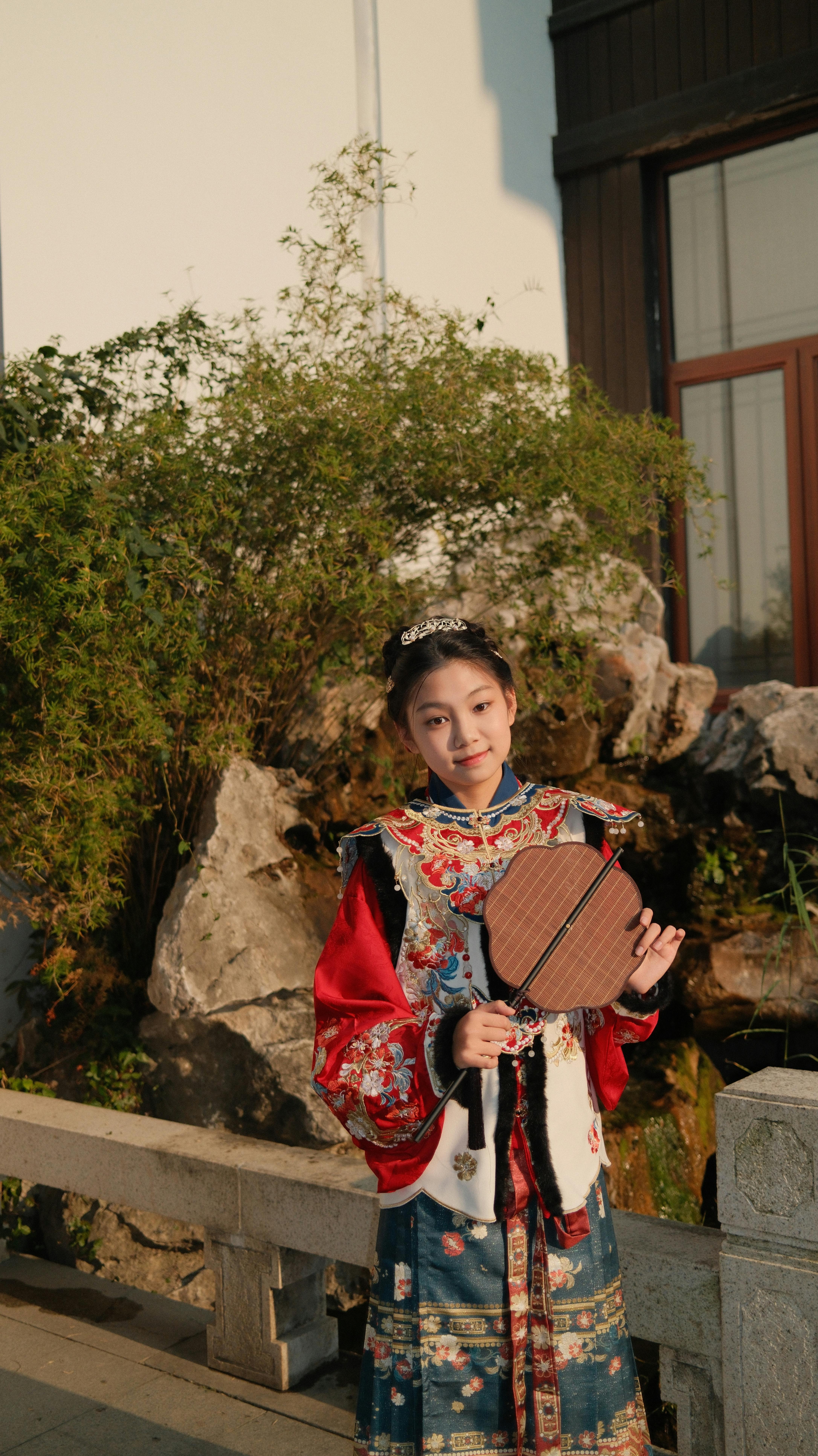 Young woman in colorful traditional dress standing outdoors holding a wooden fan.