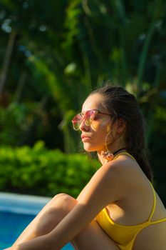 A young woman in a yellow swimsuit and sunglasses relaxes by a poolside on a sunny day.