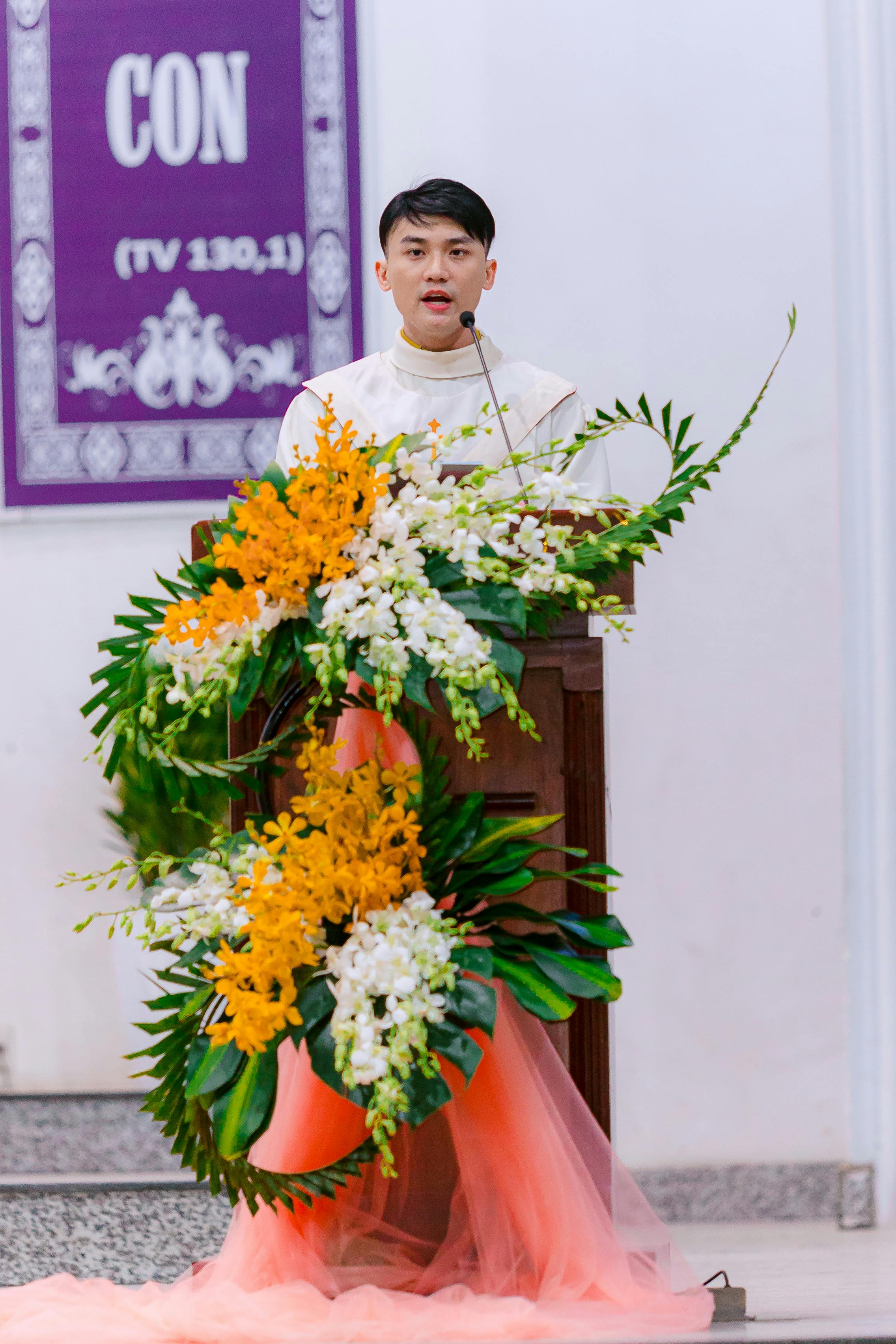Young man stands at podium adorned with vibrant flowers delivering a speech indoors.