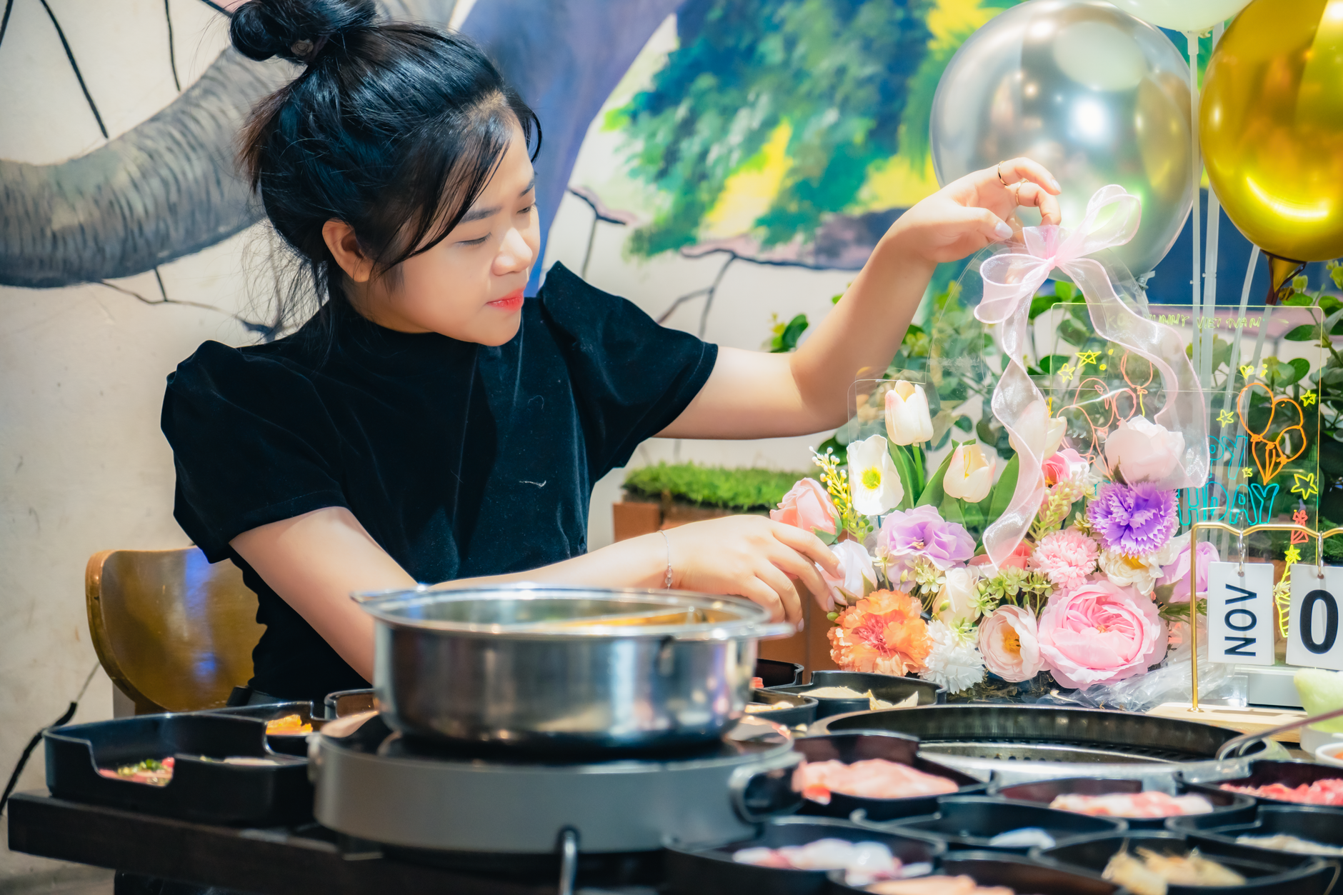 A young woman arranges flowers in a warm Vietnamese diner setting with a hotpot meal.