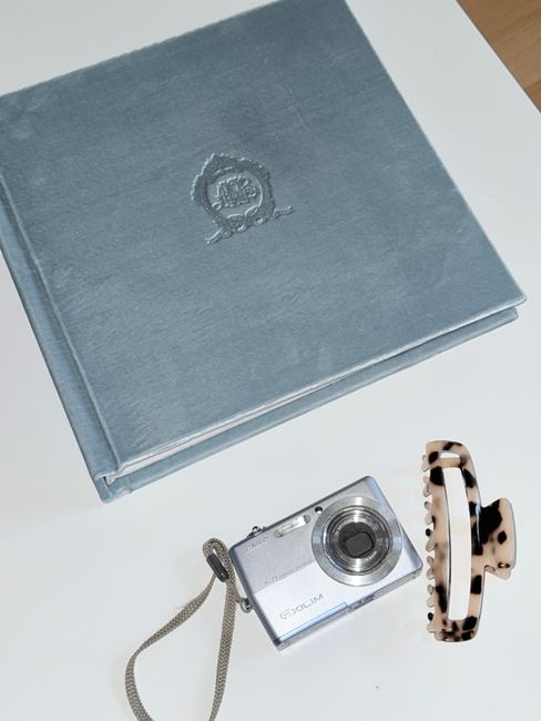 Top-down view of a vintage camera, blue photo album, and hair clip on a white surface.
