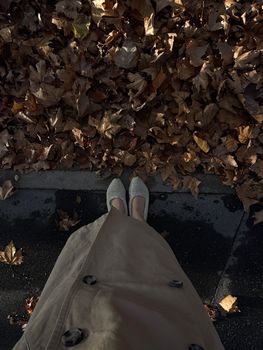 Feet in trench coat on fallen autumn leaves, borders sidewalk.