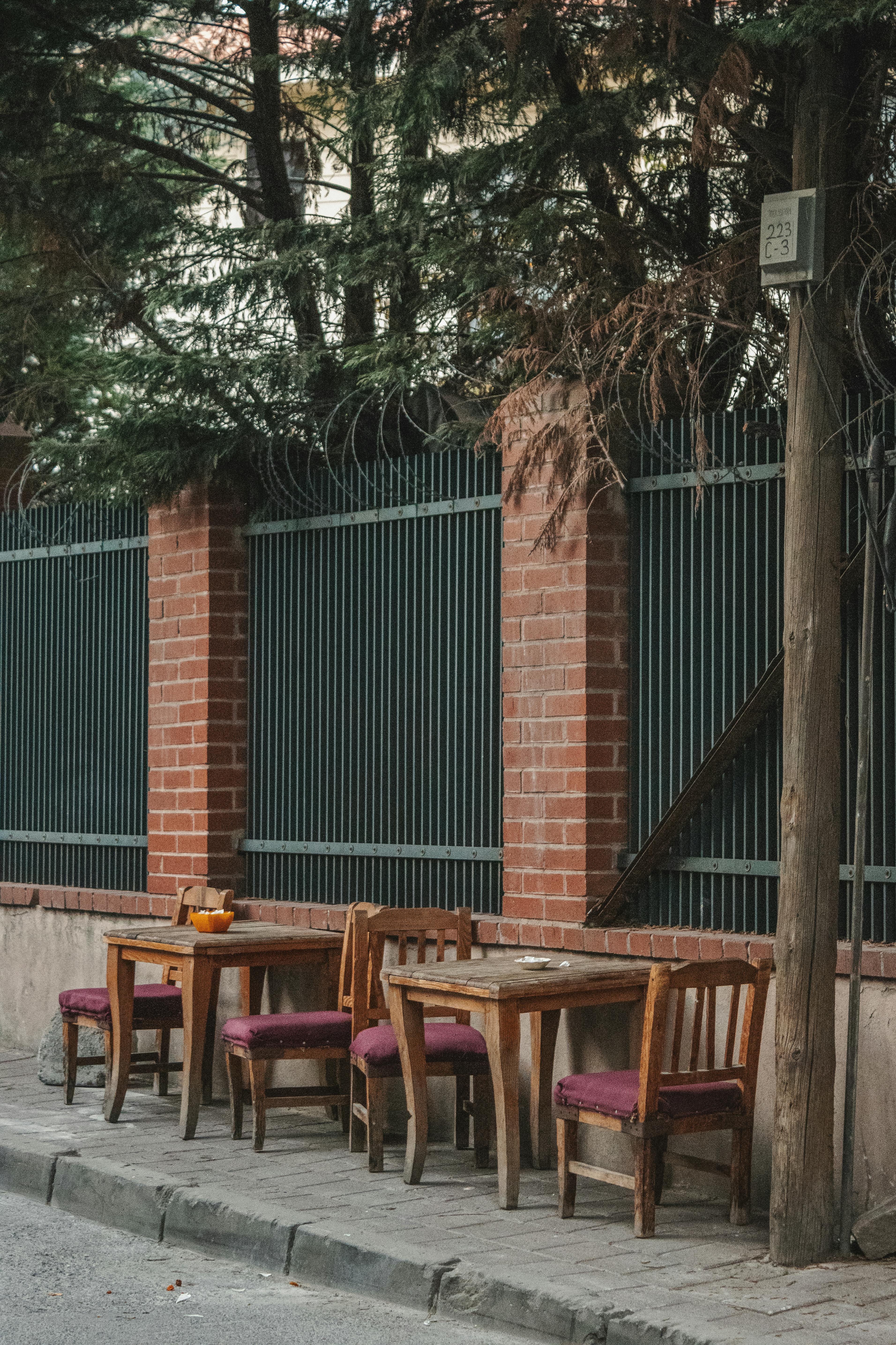 Gratis Sebuah kafe jalanan luar ruangan yang tenang dengan kursi dan meja kayu di dekat dinding bata dan pagar besi. Foto Stok