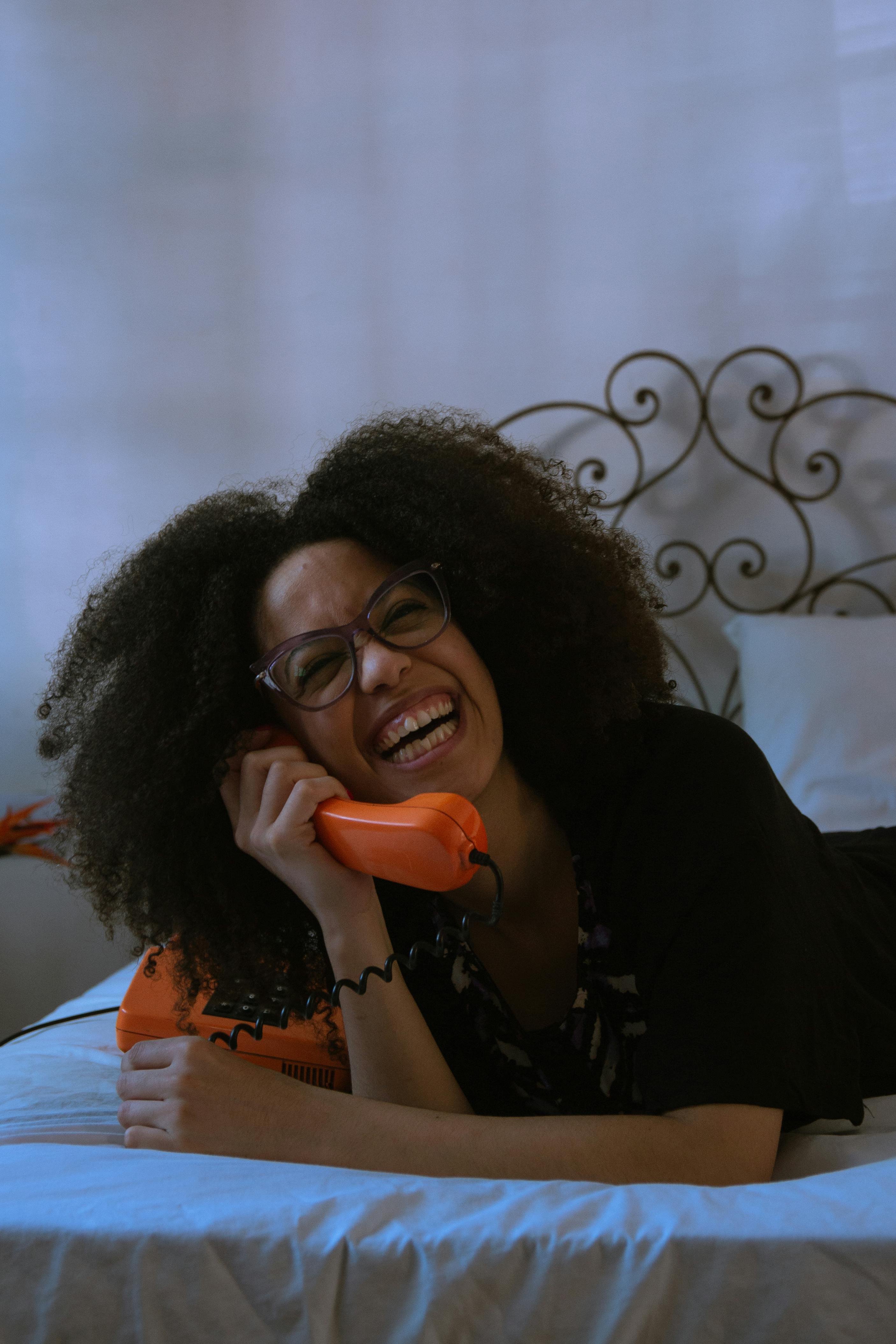 grátis Mulher de óculos rindo enquanto está deitada na cama usando um telefone de disco. Foto profissional