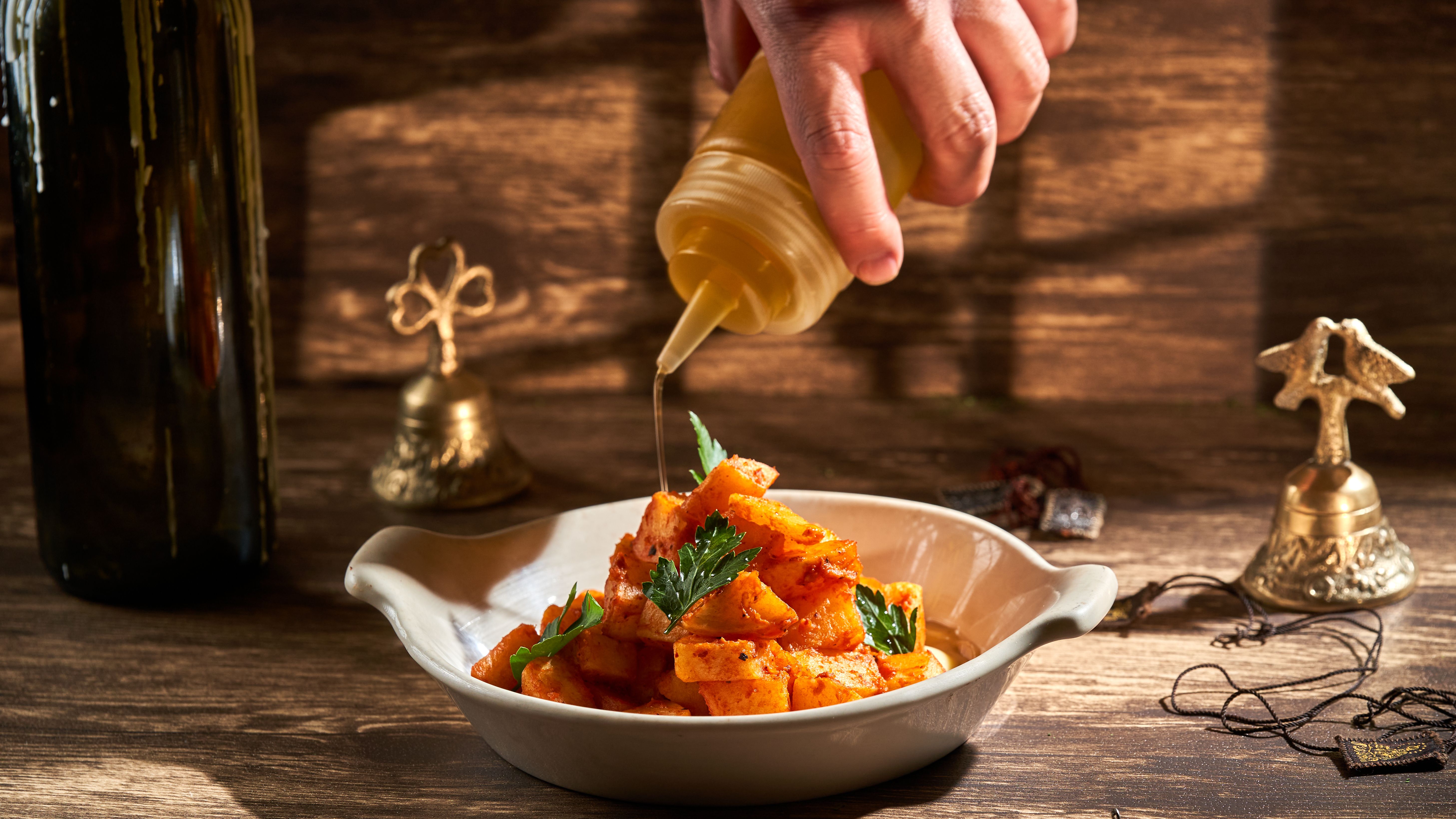 A hand drizzles oil over roasted potatoes garnished with herbs in a rustic setting.