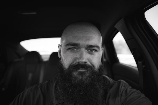 Moody black and white portrait of a man with a thick beard sitting inside a car.