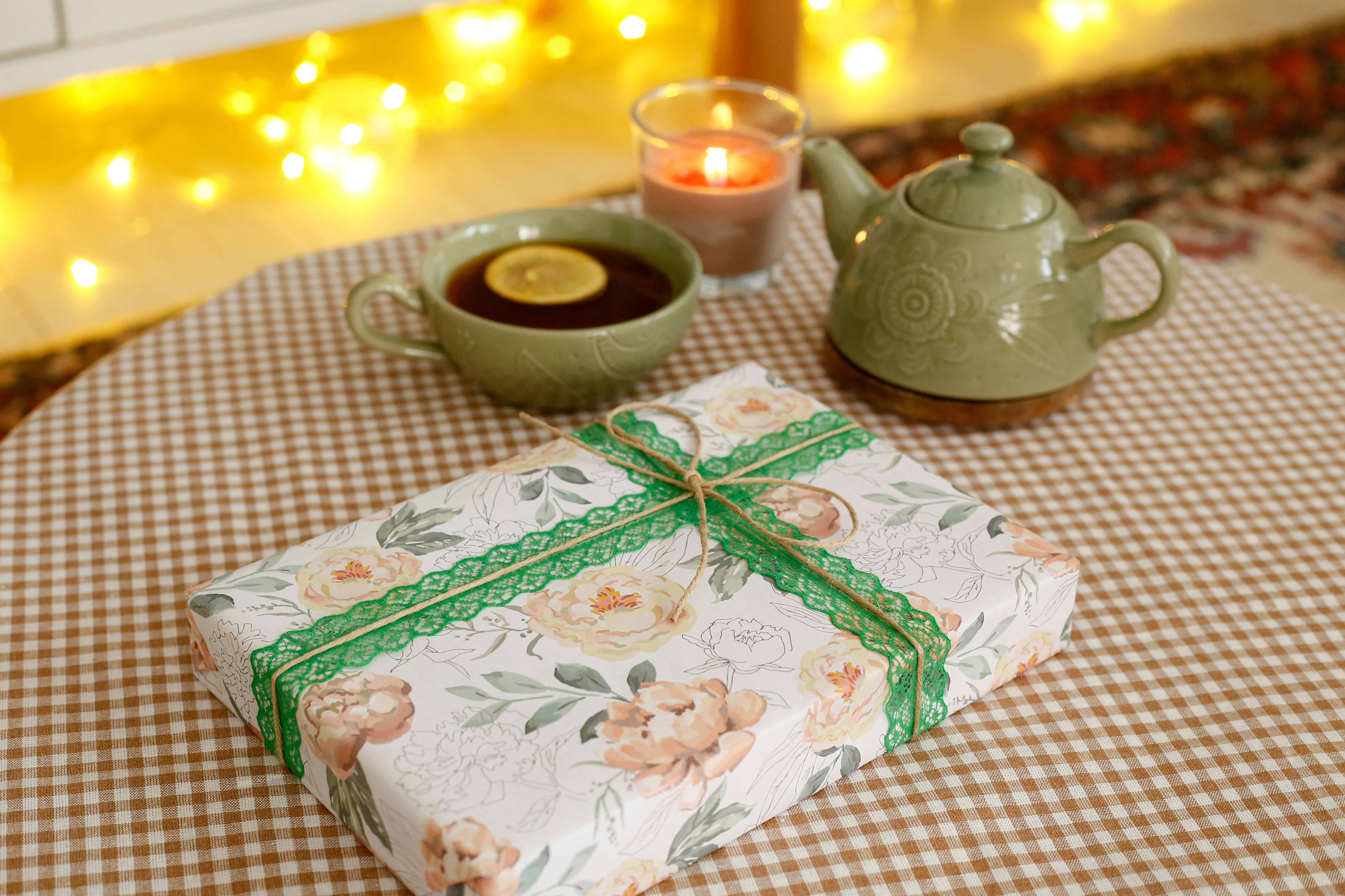 A cozy tea setting with a teapot, cup of tea, candle, and floral-wrapped gift on a checkered tablecloth.