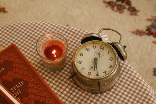 A cozy setup with a vintage alarm clock, lit candle, and patterned book on a checked tablecloth.