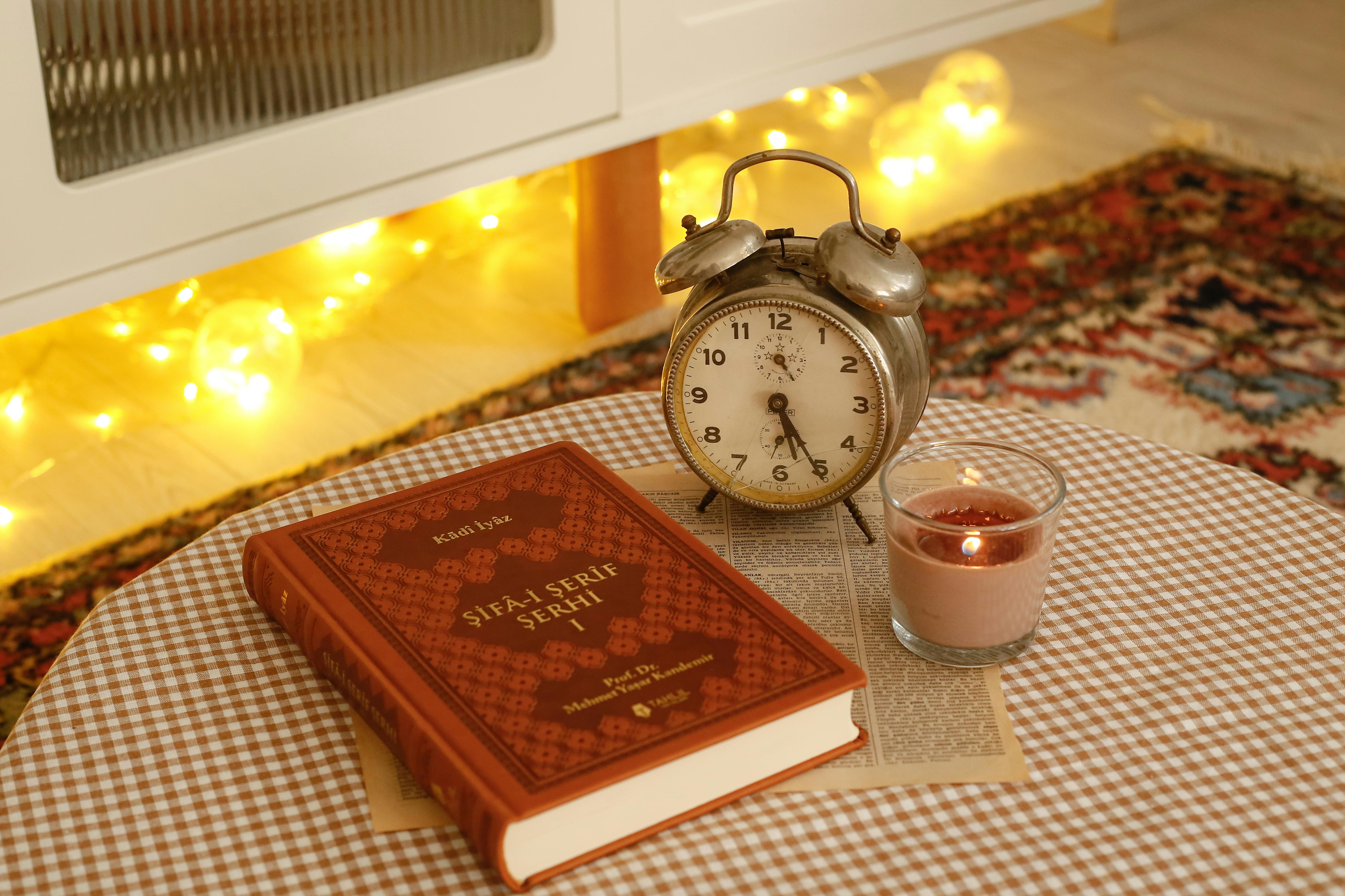 A warm, cozy still life featuring a vintage clock, book, and candle on a checkered tablecloth.