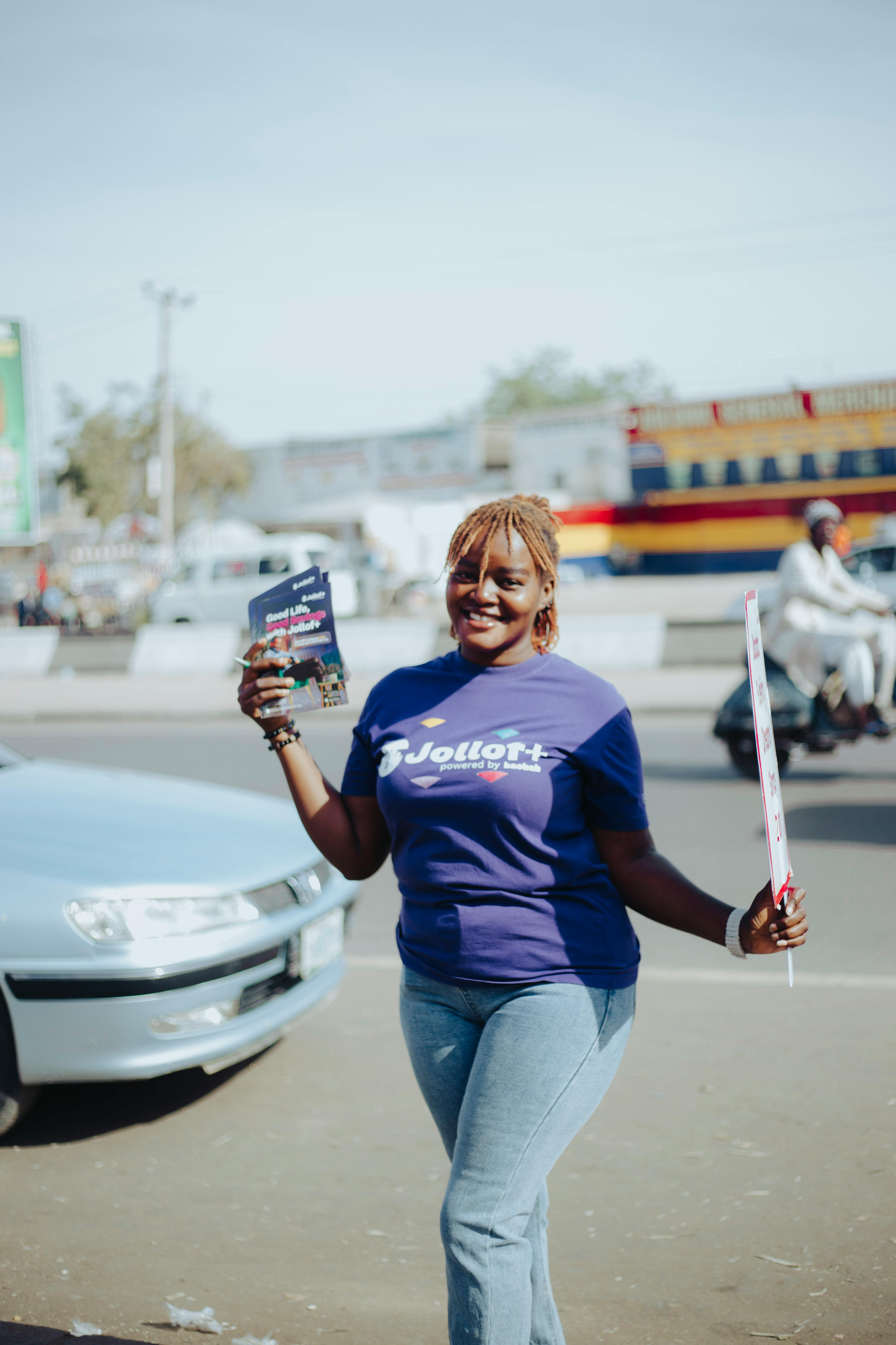 Cheerful woman wearing a purple T-shirt and jeans, holding flyers on a sunny day.
