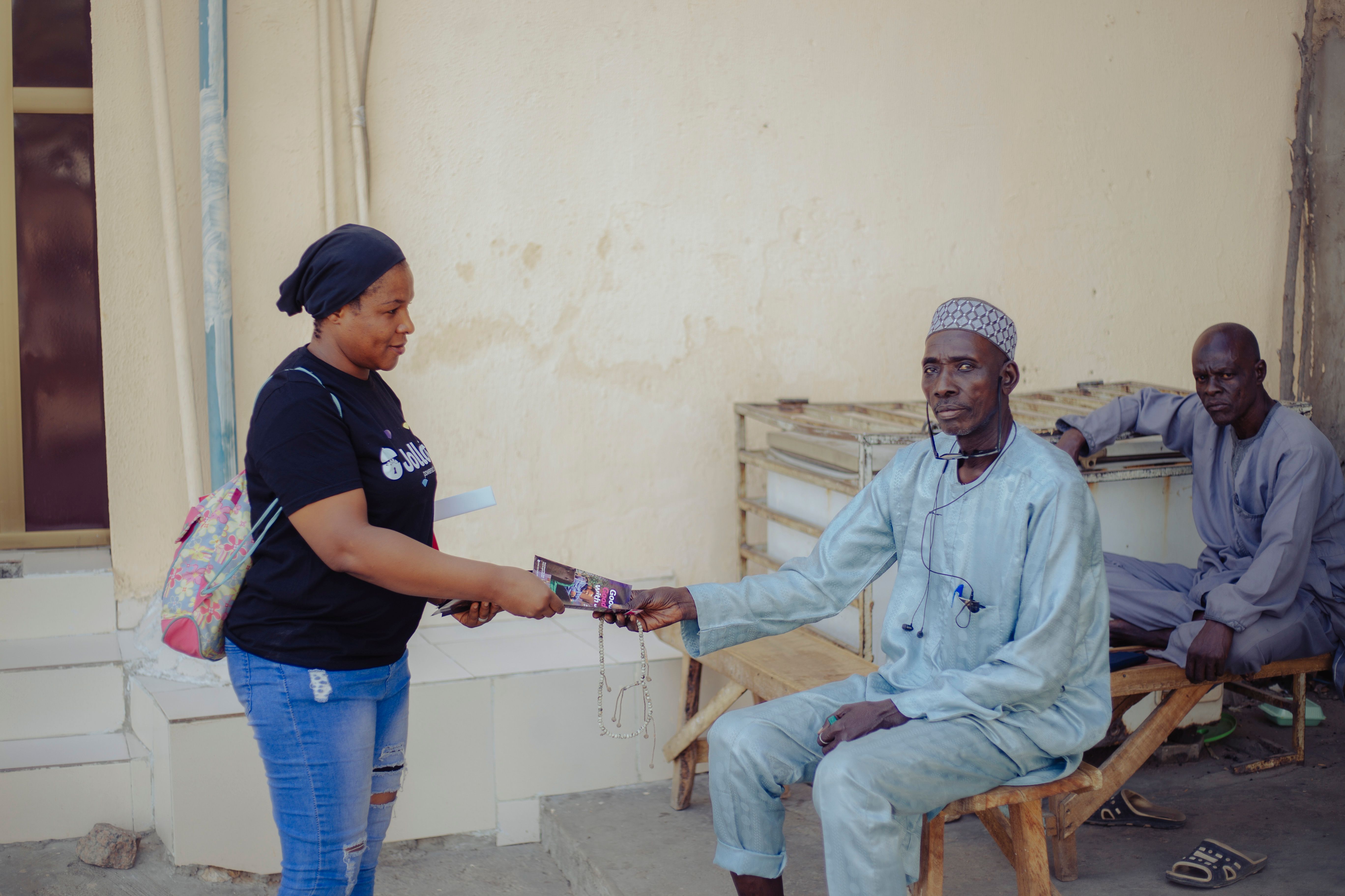 A woman in casual attire gives an item to a seated man in a relaxed outdoor setting.