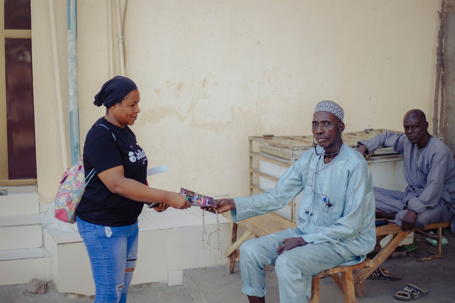 A woman in casual attire gives an item to a seated man in a relaxed outdoor setting.