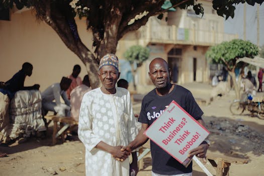 Two African men shaking hands outdoors with a business sign, community setting.