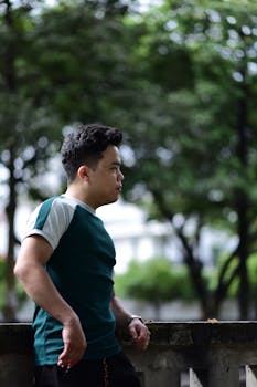 A young man in a turquoise shirt leans on a stone wall in a green park.