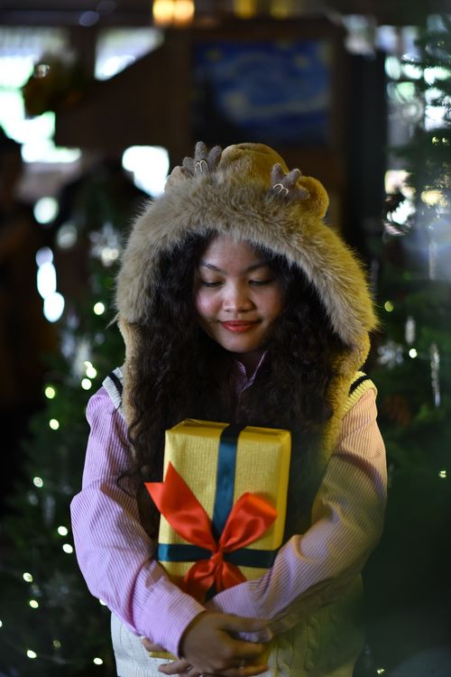 Free A woman in festive winter attire holds a wrapped gift surrounded by holiday lights. Stock Photo