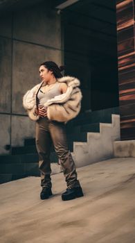 Fashionable woman posing in a fur coat and boots on modern concrete stairs.
