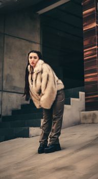 A stylish woman poses in a fur coat and trousers on modern urban stairs, showcasing contemporary winter fashion.