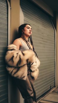 Stylish woman in fur coat and braids leans against a city wall, capturing urban fashion vibes.