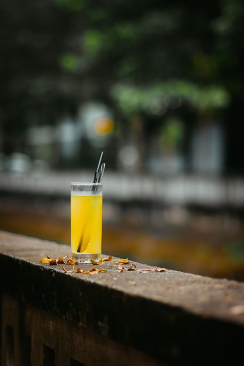 Free A glass of bright yellow juice with a straw on an outdoor ledge, surrounded by urban scenery. Stock Photo
