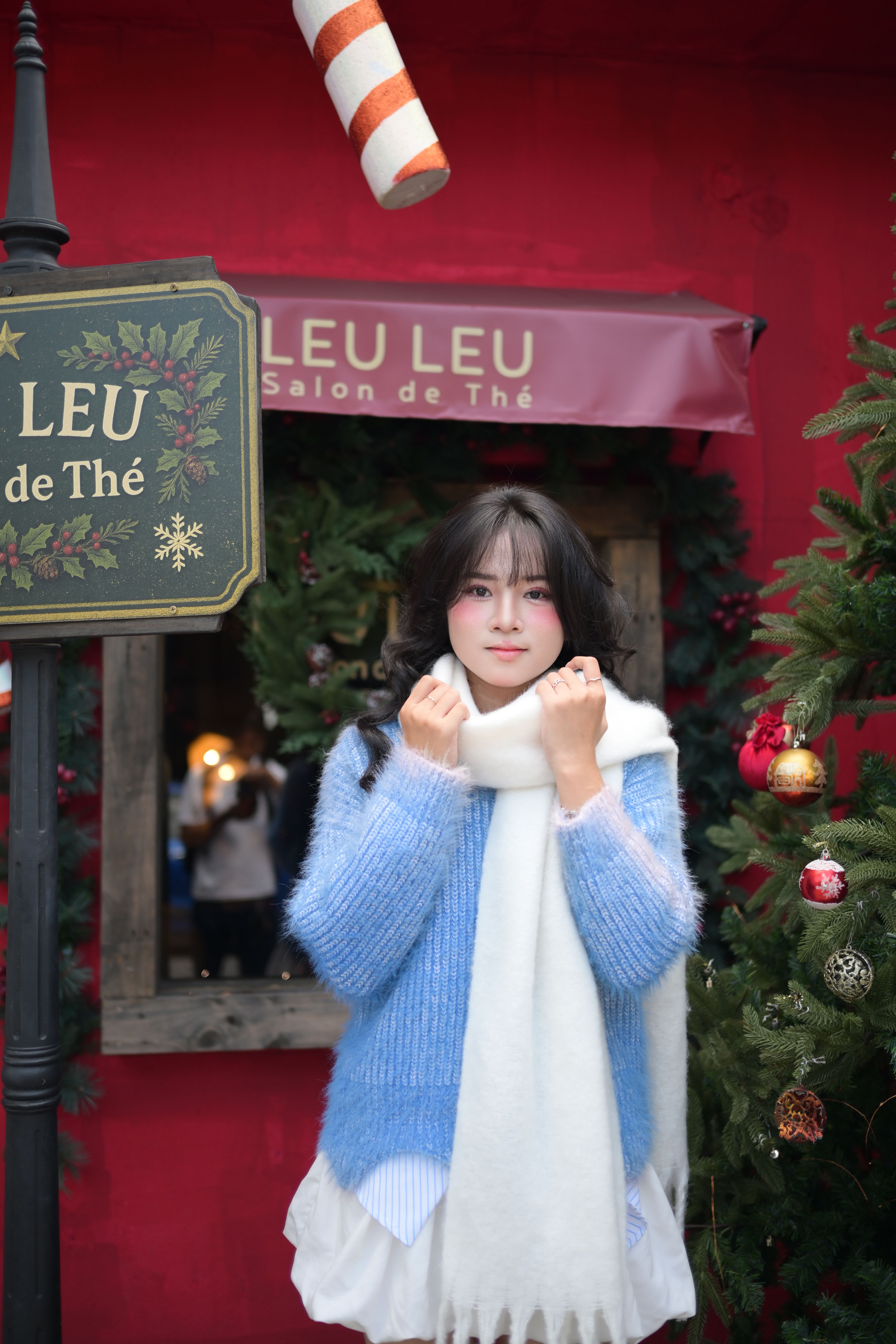 Young woman wearing blue sweater and scarf outside decorated cafe during Christmas season.