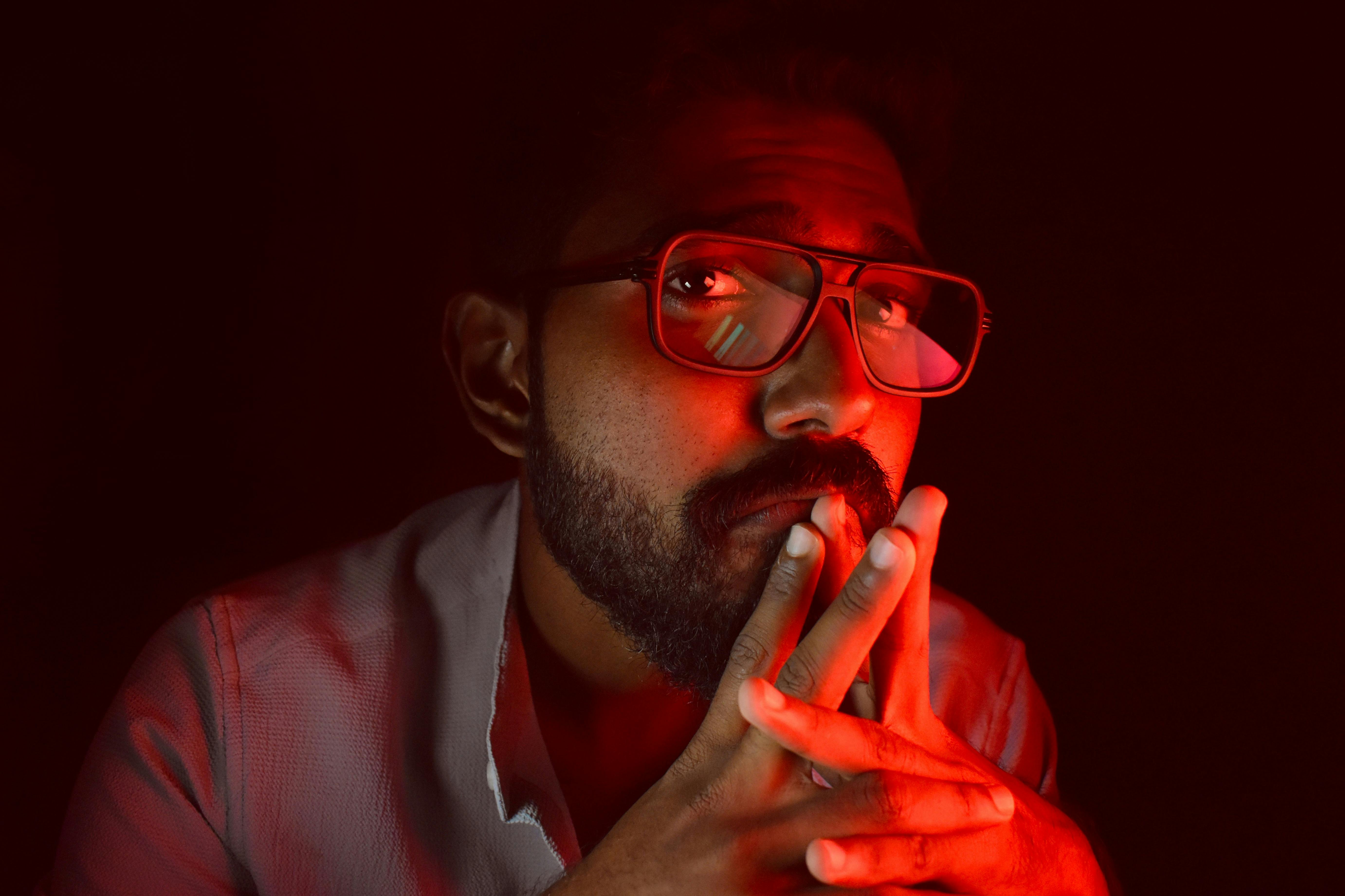 Gratis Un uomo pensieroso, con una luce ambientale rossa, occhiali e barba, che guarda verso l'alto. Foto a disposizione