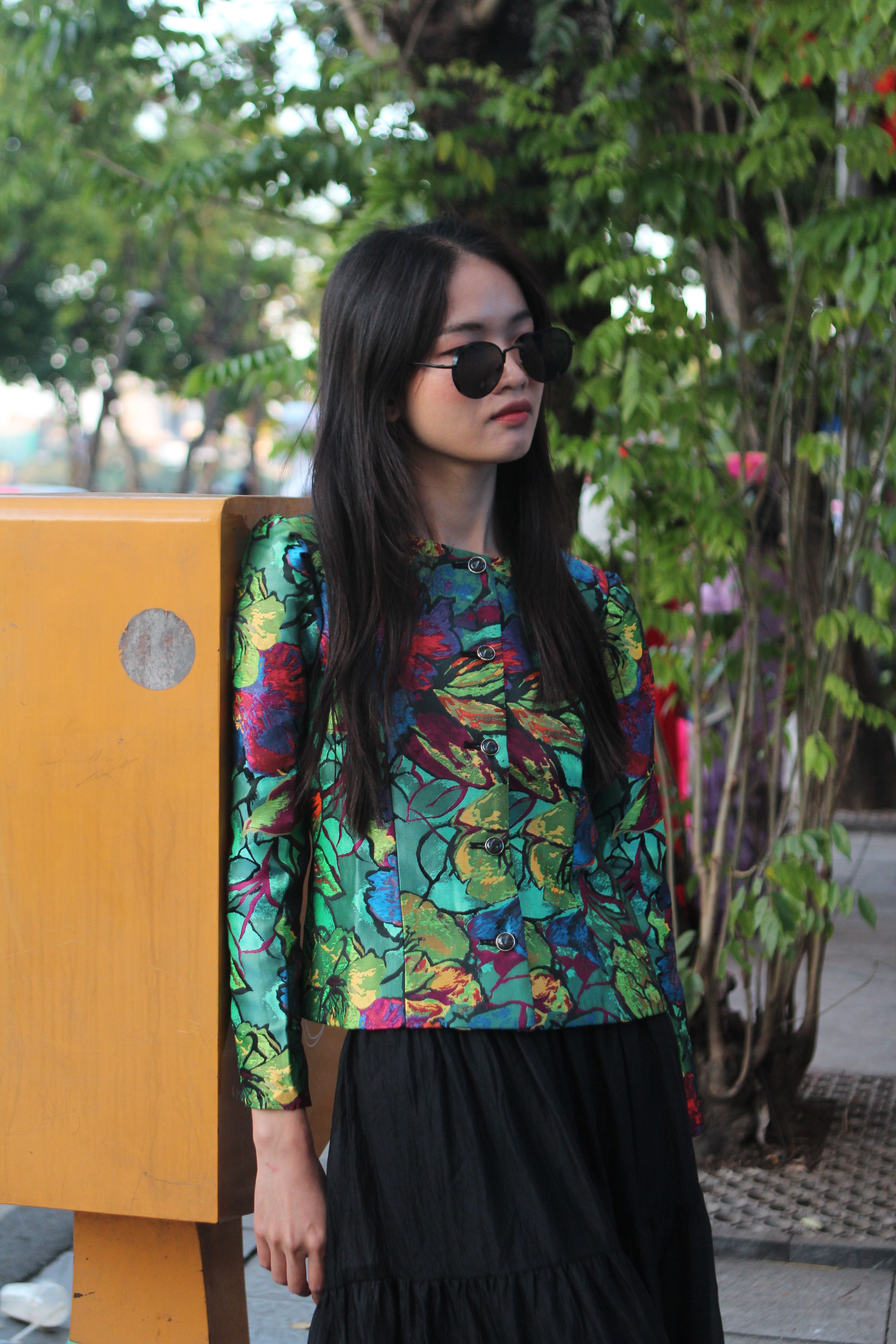 Stylish young woman in colorful floral jacket and sunglasses standing outdoors.