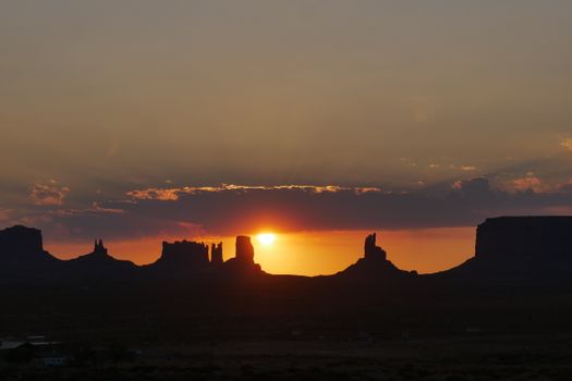 Silhouette of Monument Valley at sunset with vivid orange sky and sun peeking through rock formations.