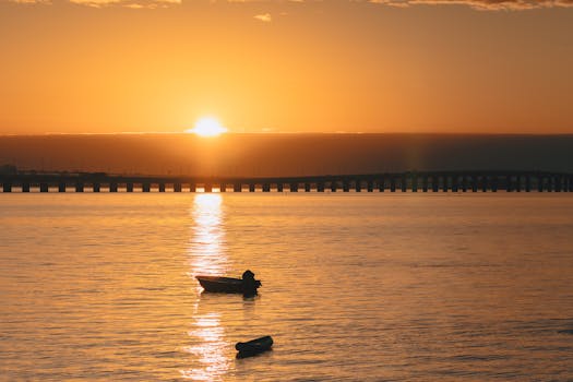 Serene sunrise over ocean with silhouetted boats and bridge, creating a peaceful ambiance.