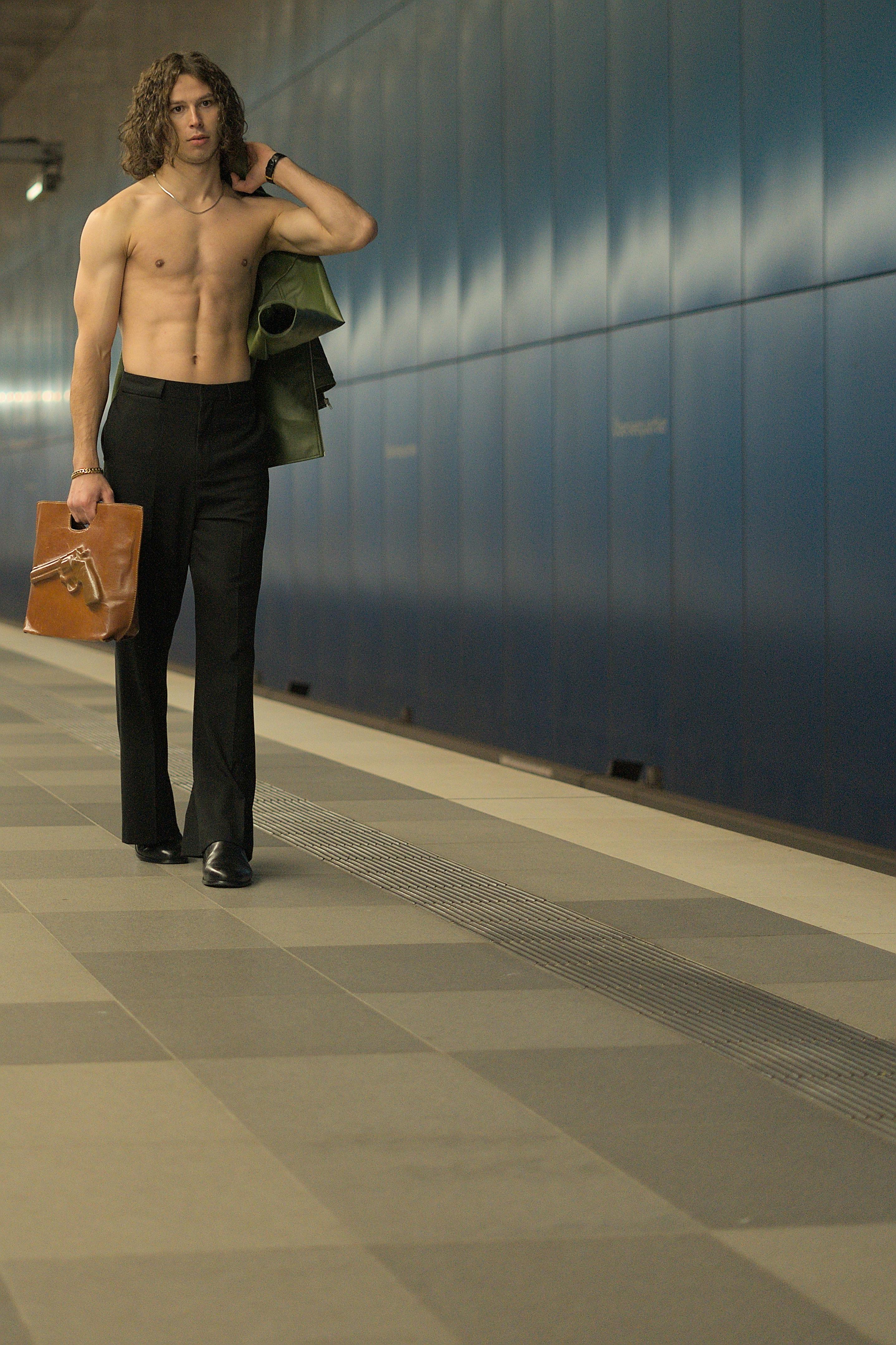 A muscular man walks confidently through a subway corridor carrying clothes and a briefcase.