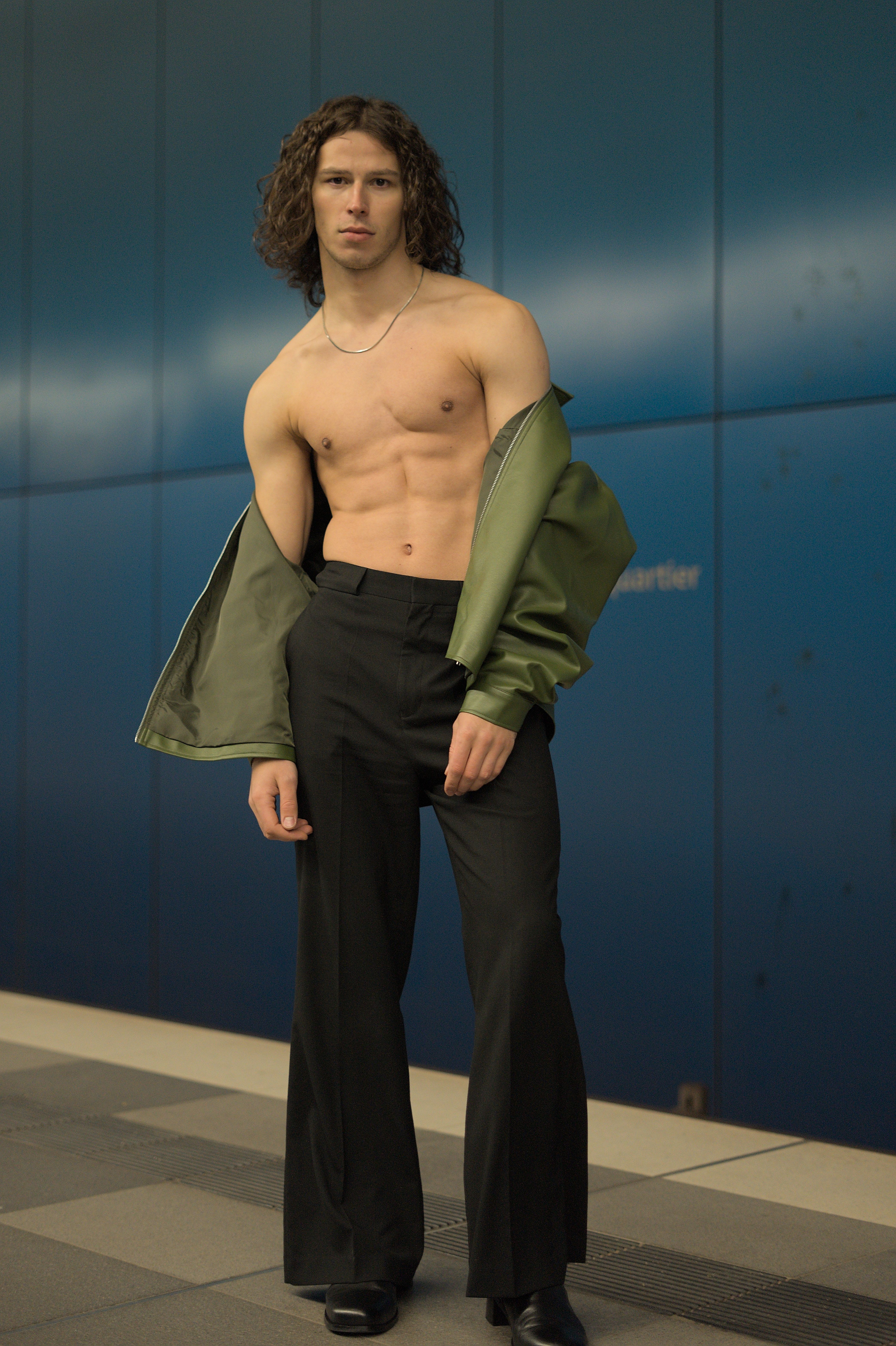Shirtless man wearing wide-leg pants and green jacket in urban setting. Modern fashion style.