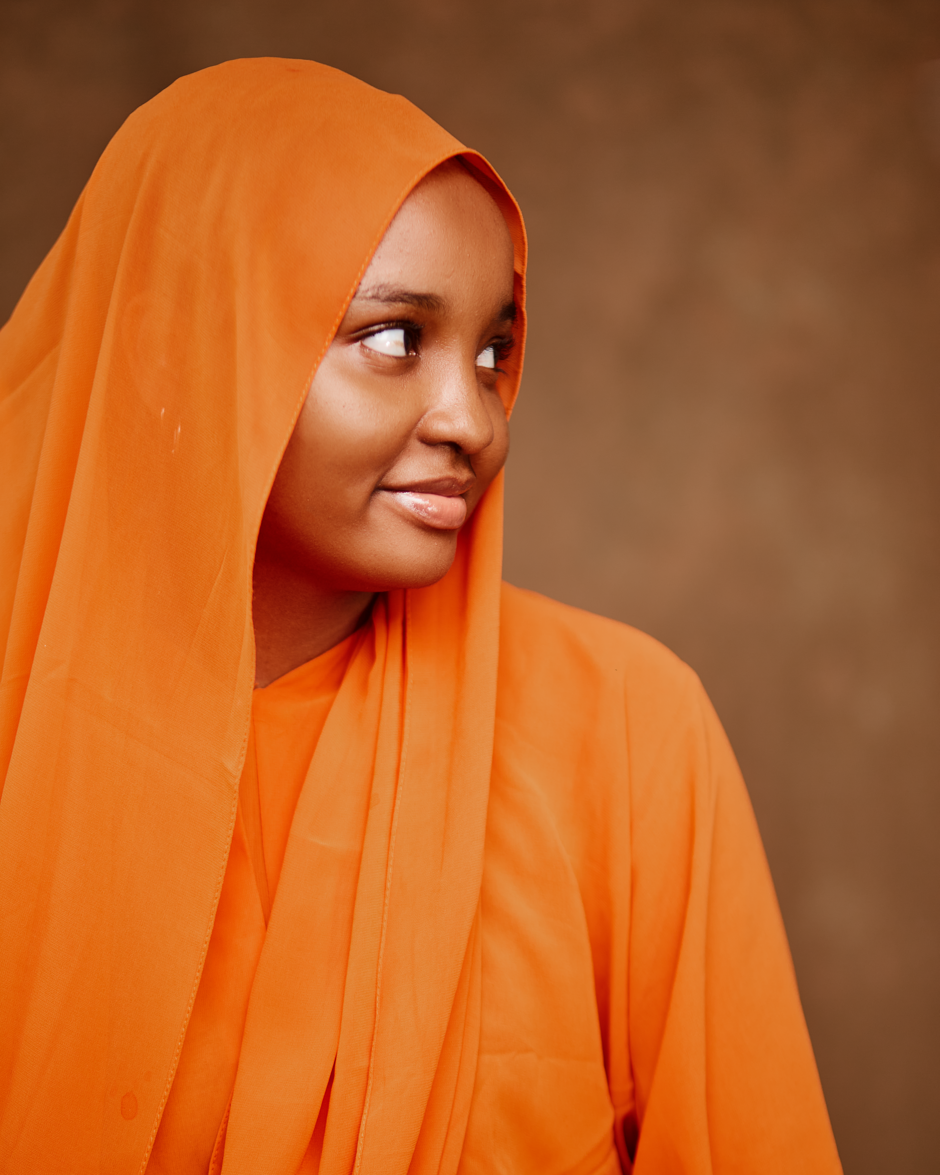 Young woman in orange hijab against brown background looking to the side.