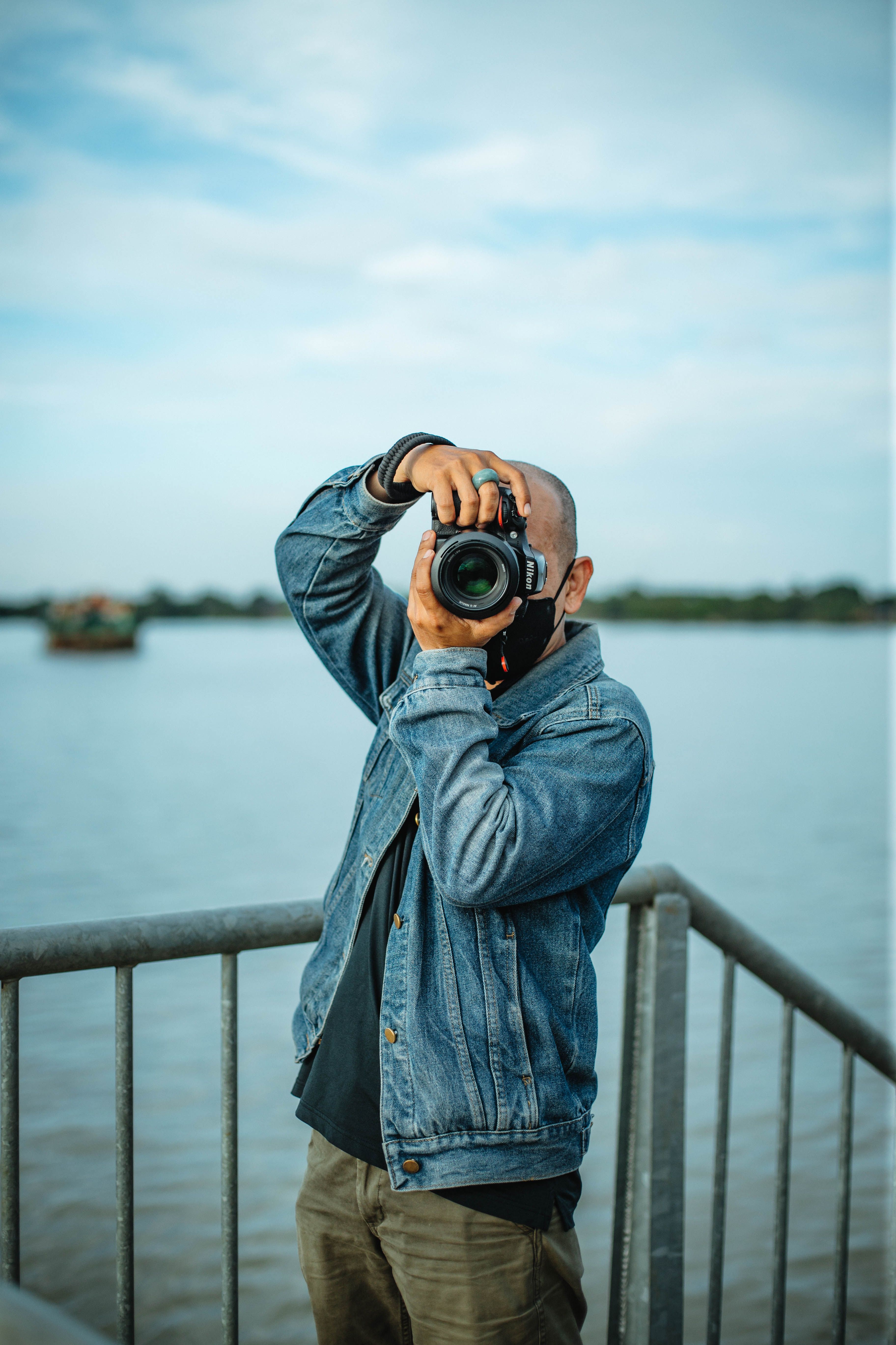 Man with camera taking photos by a river, wearing denim jacket outdoors.