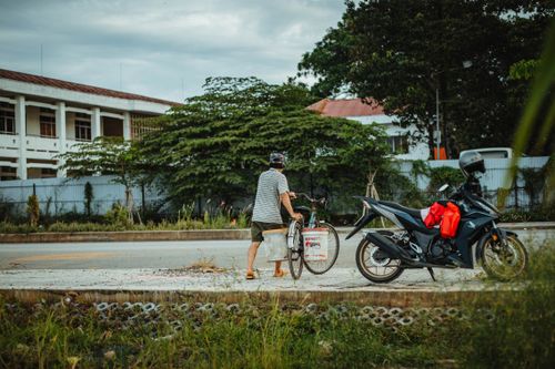 Free An adult man walking a bicycle near a parked motorcycle on a street with trees and buildings. Stock Photo