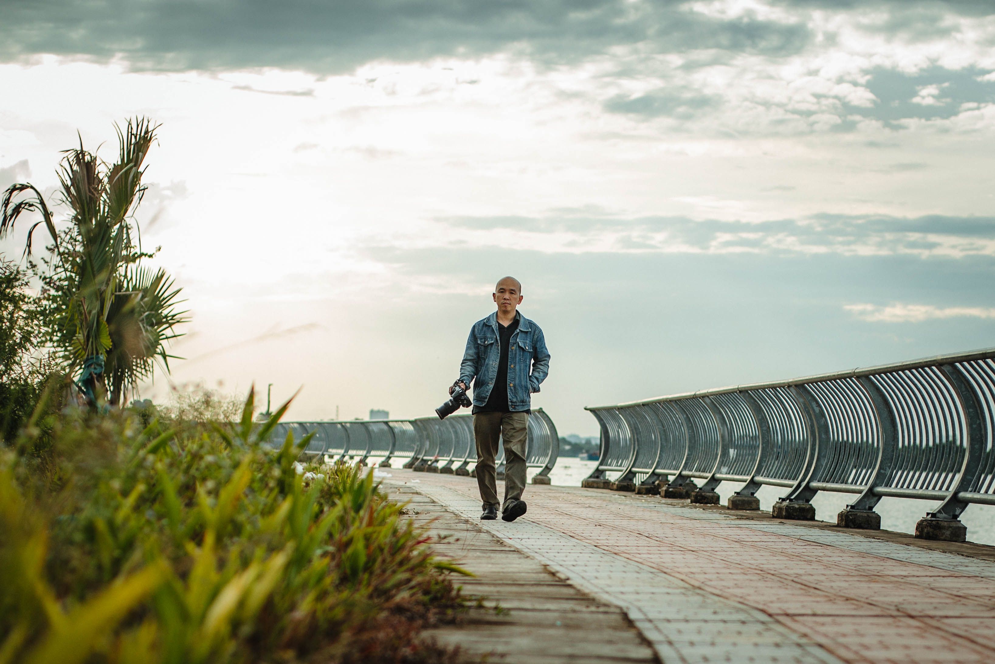 A man with a camera walks along a scenic riverside path during the day.