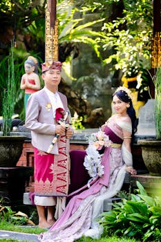 A couple in traditional Balinese attire pose outdoors in lush greenery.