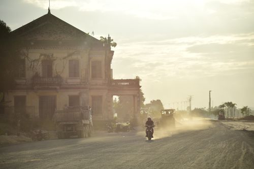 Free A motorcycle rides on a dusty road beside an old, abandoned building in warm light. Stock Photo
