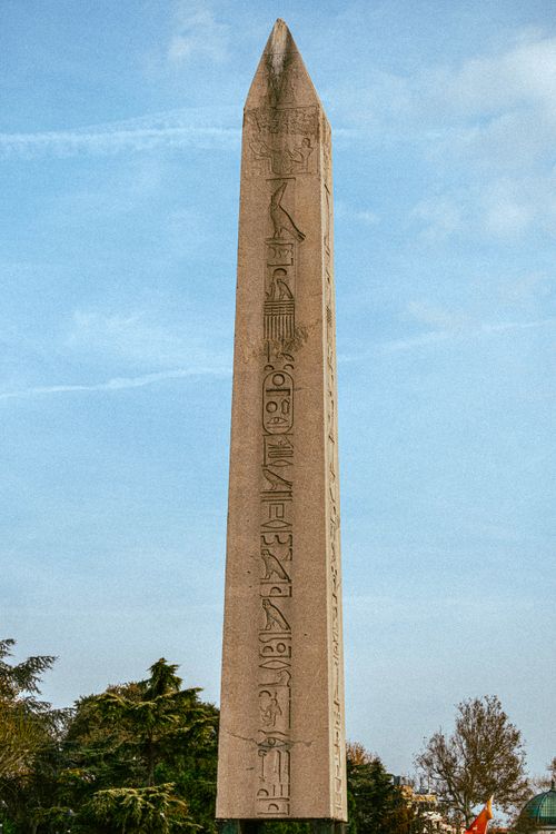 Free Tall ancient obelisk with hieroglyphs stands against a clear blue sky with trees below. Stock Photo
