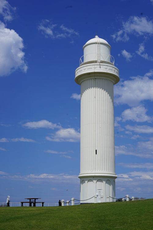 Free Tall white lighthouse standing against a vibrant blue sky with clouds. Stock Photo