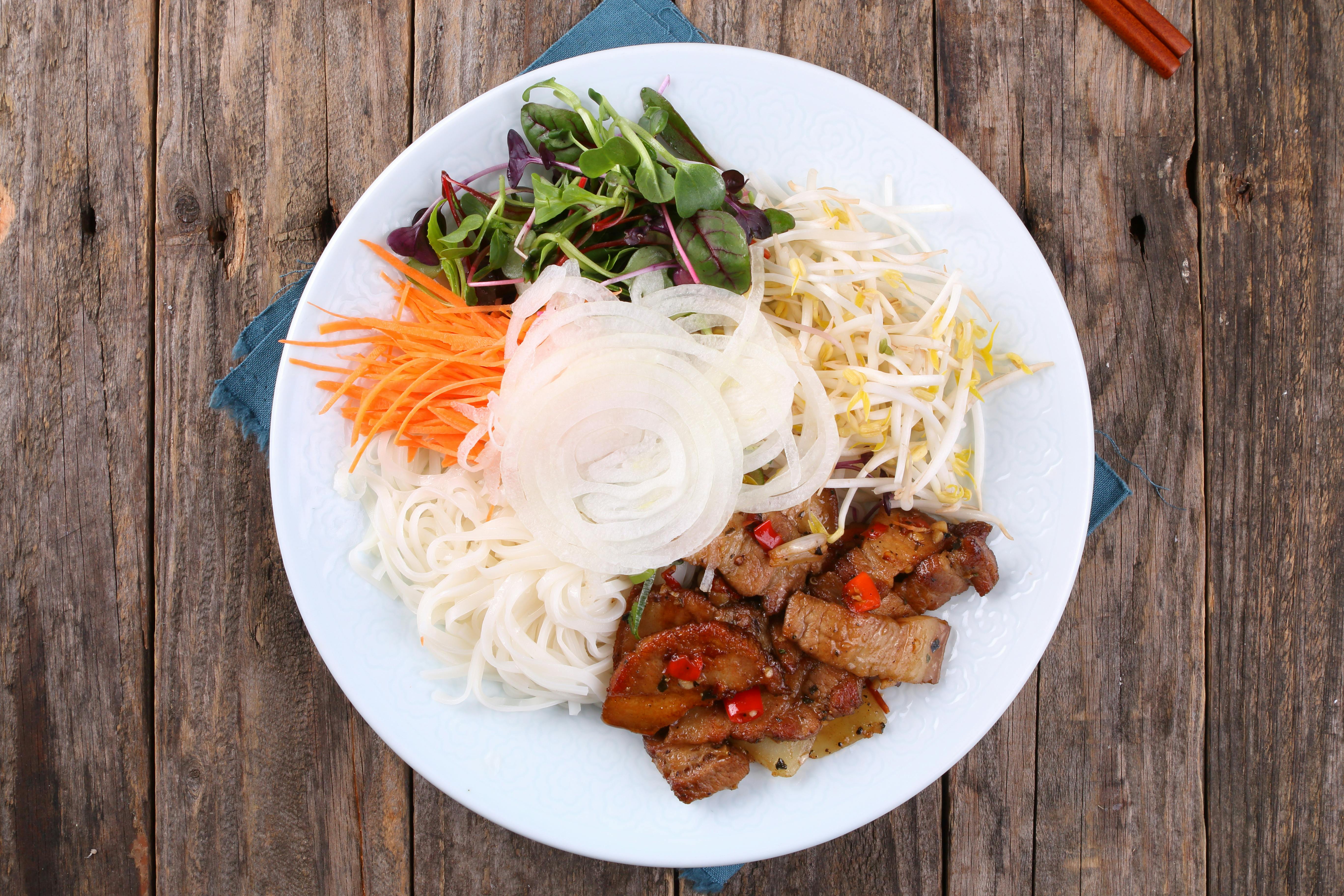 A vibrant Vietnamese noodle salad with pork, vegetables, and sprouts on a wooden table.
