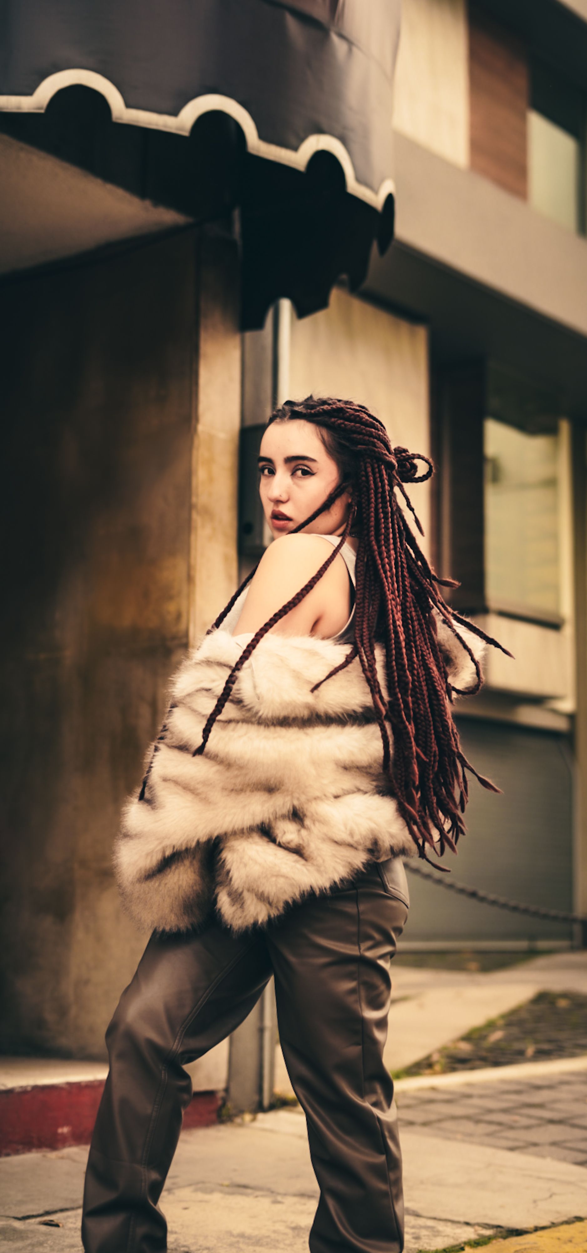 Mulher fashion com tranças longas em casaco de pele posa ao ar livre em cenário urbano.