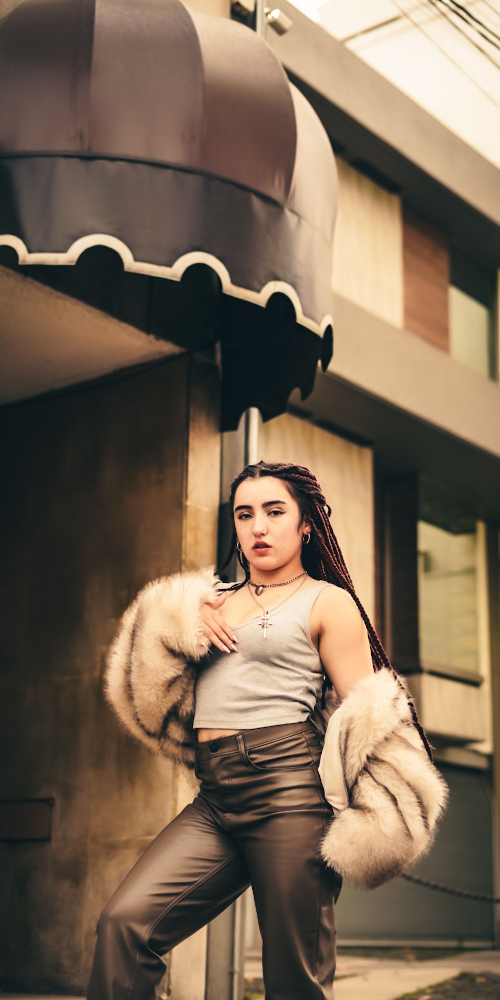 Mulher elegante com casaco de pele posa com confiança sob a marquise de um prédio em ambiente urbano.