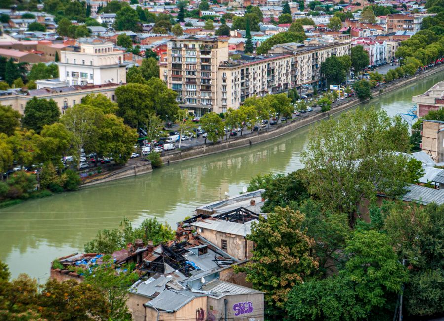 Aerial view of city buildings lining a river surrounded by lush trees and urban scenery.
