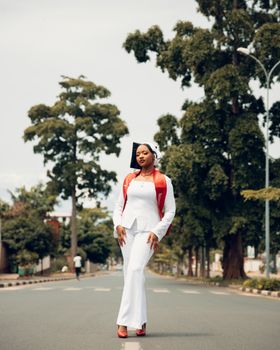 Elegant woman in white and red attire walks confidently on a tree-lined street in Burundi.