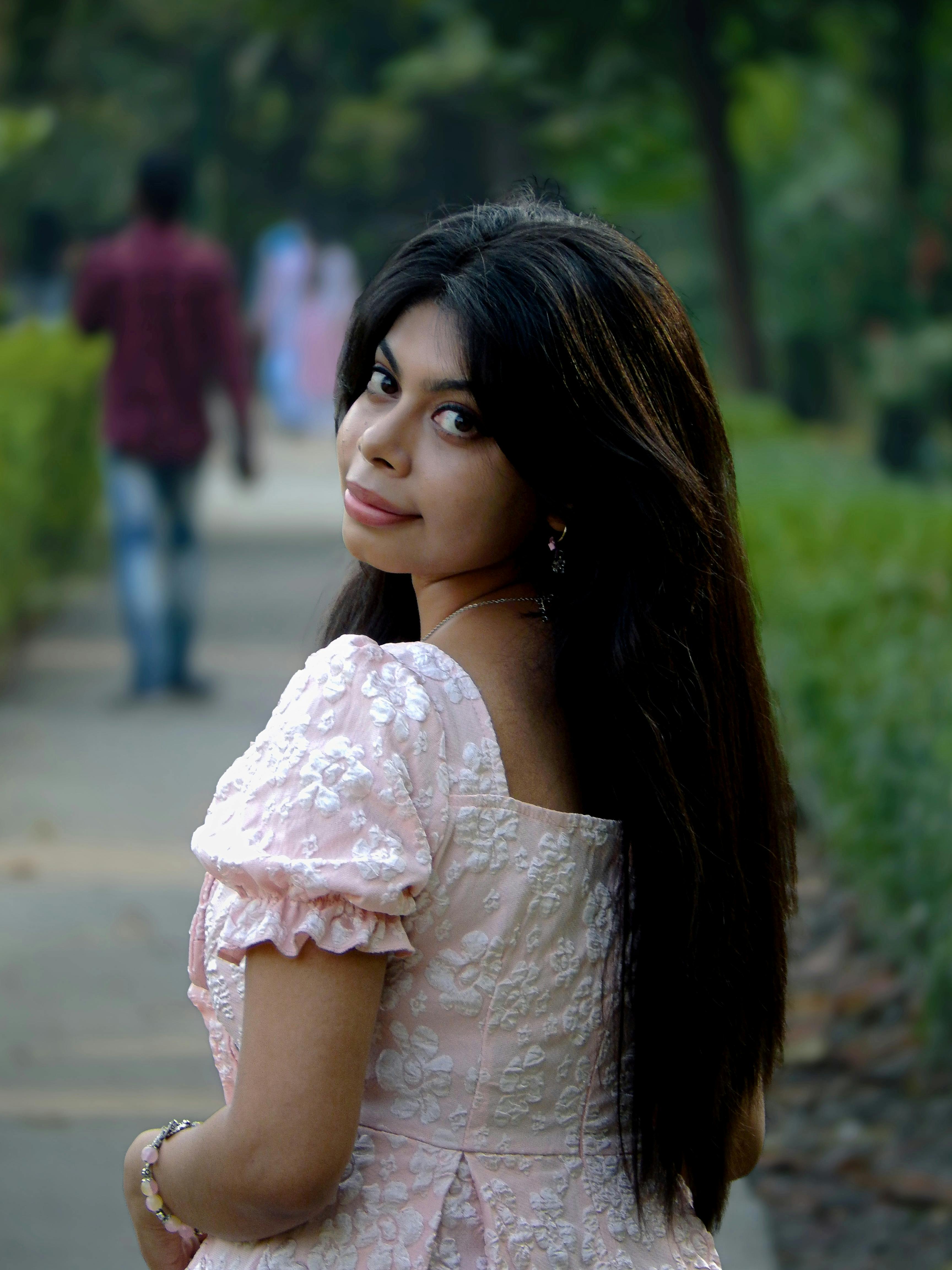 A young woman in a pink dress poses outdoors in an Indian garden, exuding elegance and charm.