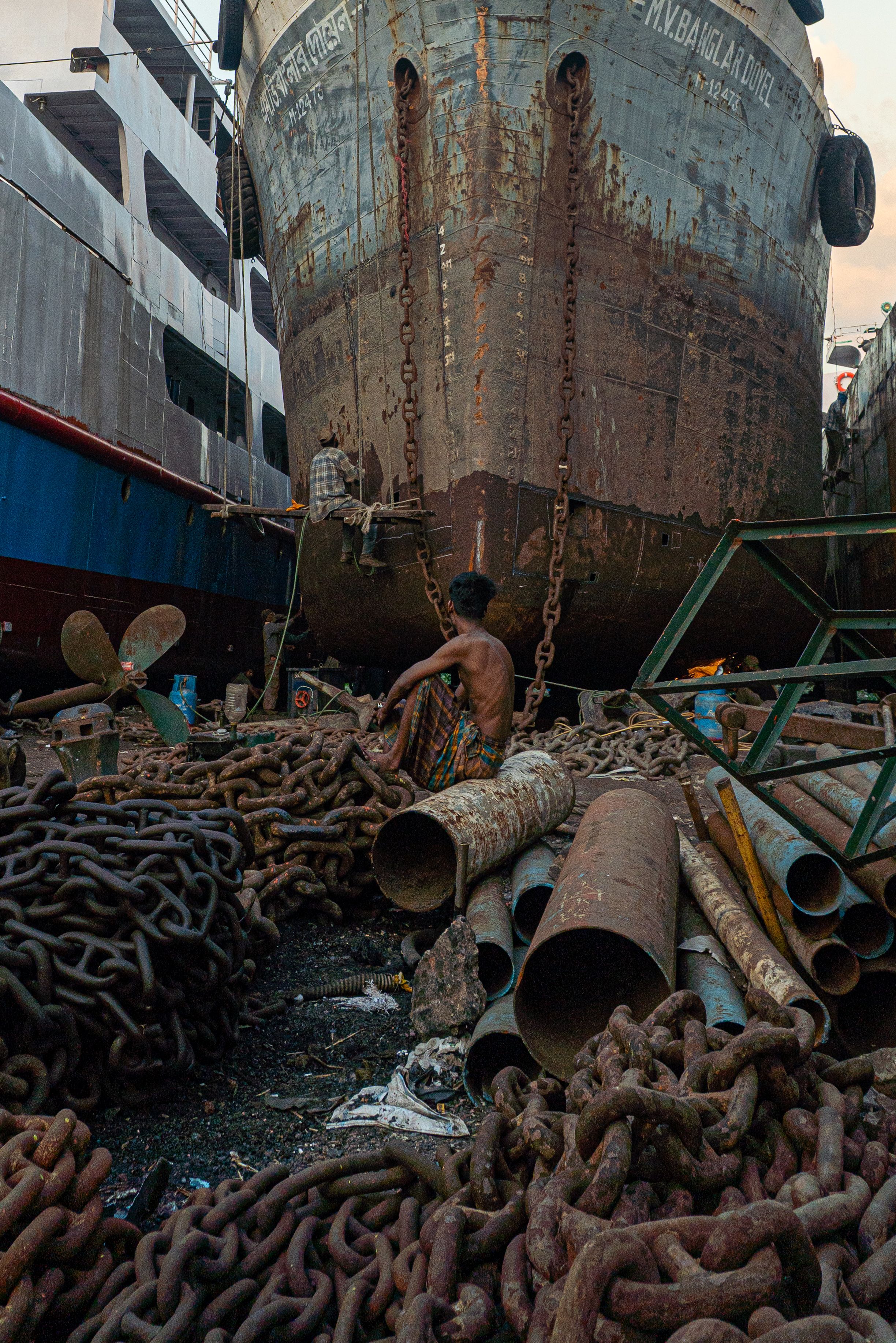A man sits in a shipyard surrounded by rusty chains in Dhaka, Bangladesh, showcasing daily life.